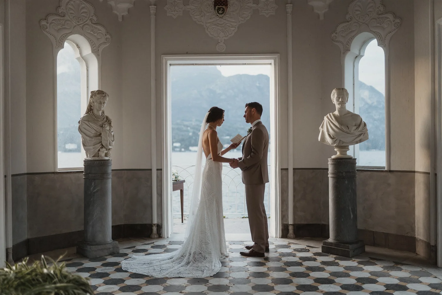 Bride and groom holding hands during their wedding ceremony in an elegant indoor setting with classical sculptures on pedestals, large windows revealing mountains in the background, and decorative architectural details.