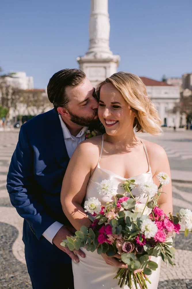 A newlywed couple outdoors with a city monument in the background. The groom is kissing the bride on her cheek, who is smiling and holding a bouquet of flowers.