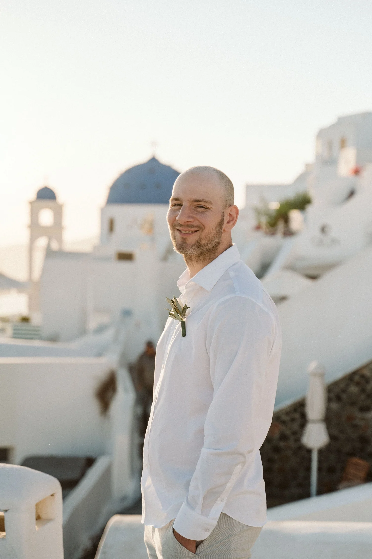 A smiling man with a shaved head wearing a white shirt and light-colored pants, standing outdoors with white buildings and boats in the background, near a body of water during daylight.