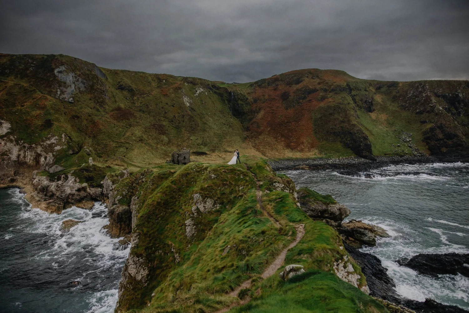 A rugged coastline with green cliffs, a narrow path, and a stormy sky, featuring a couple dressed in wedding attire standing near the edge of a rock formation.