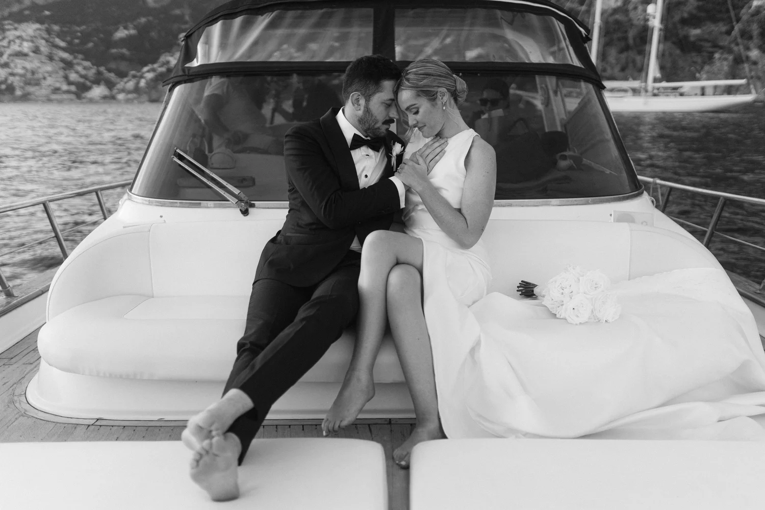 Black and white photo of a bride and groom sitting closely together on a boat, holding hands and touching foreheads with eyes closed, wedding bouquet on the bench in front of them.