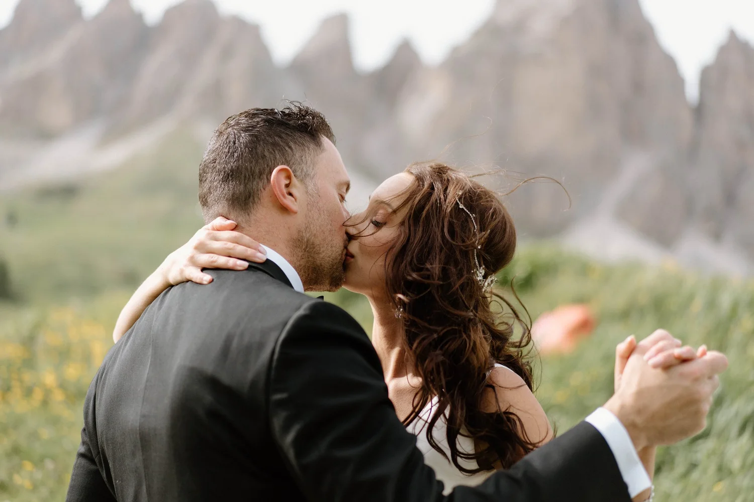 A couple sharing a kiss outdoors, dressed formally, possibly at a wedding, with a scenic mountainous background.