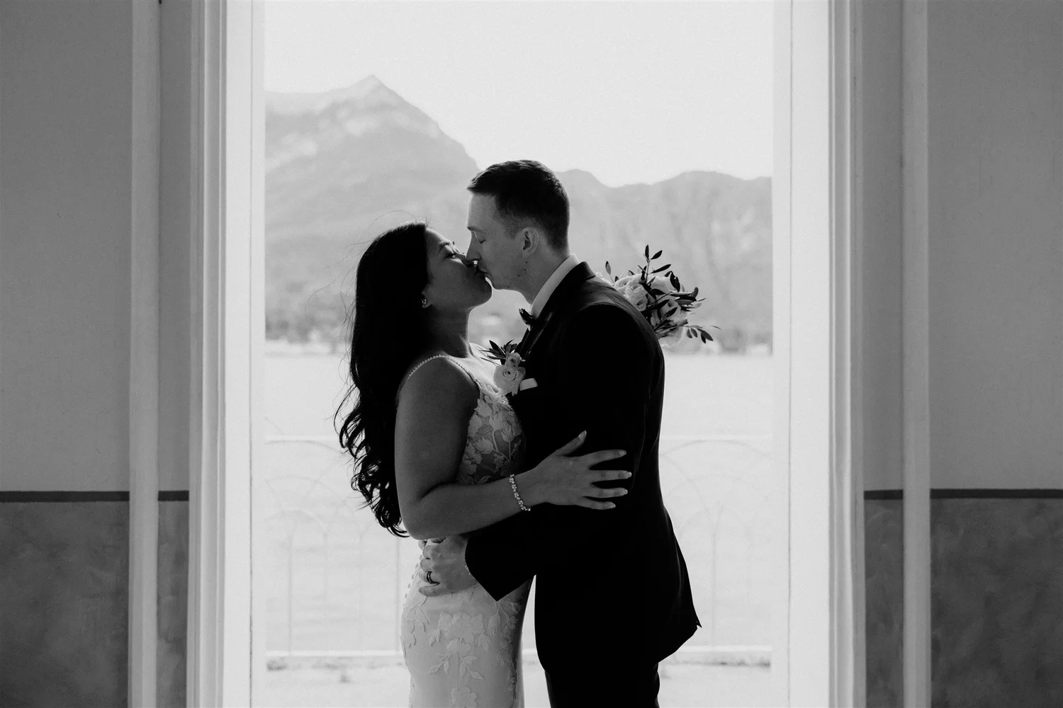A black and white photo of a bride and groom sharing a kiss in front of a window with scenic mountains in the background.