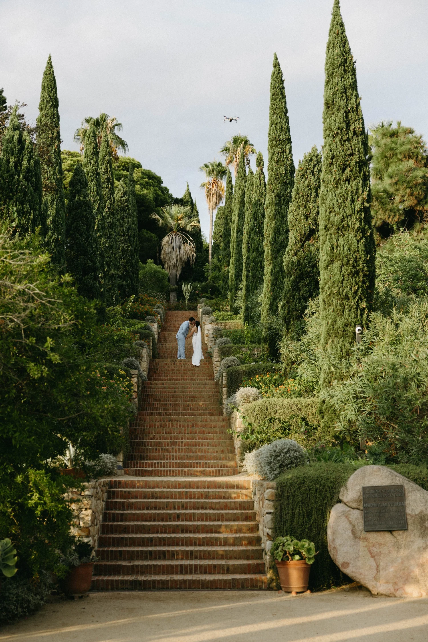 A couple dressed in wedding clothes sharing a kiss on a staircase in a lush garden with tall cypress trees and palm trees. The staircase is made of brick, and there are plants and rocks on either side, with a drone in the sky above.
