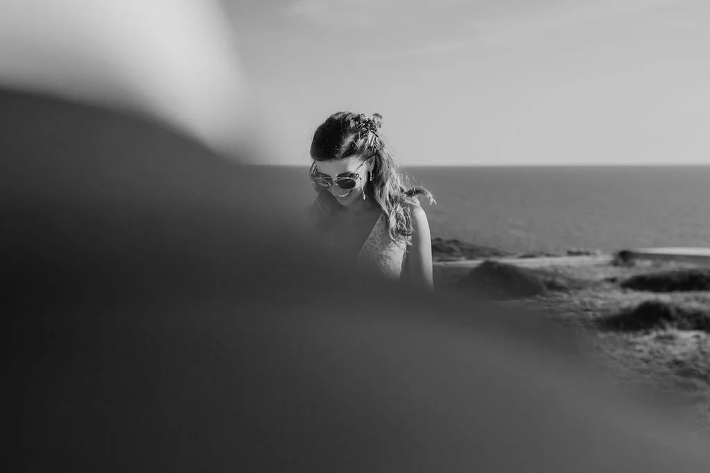 A woman with sunglasses smiling outdoors near the beach, with rocks and ocean in the background, partially obscured by a blurred foreground.