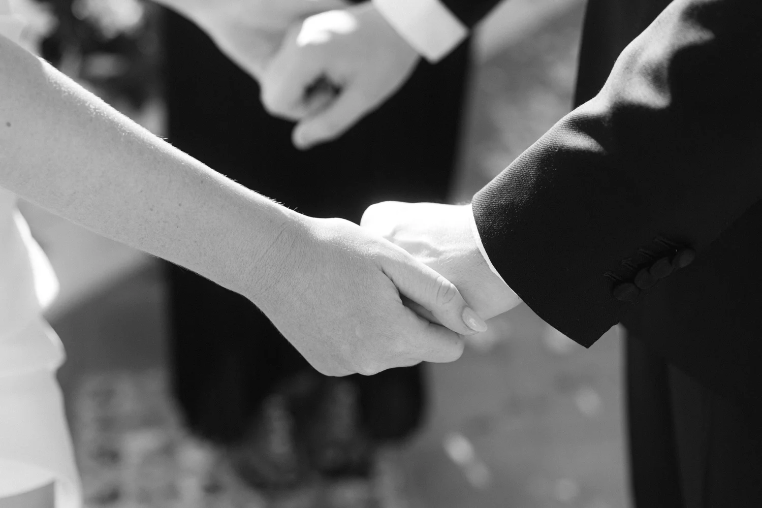 Close-up of two people holding hands, one in a dark suit and the other in a light-colored dress, in a black-and-white photograph.