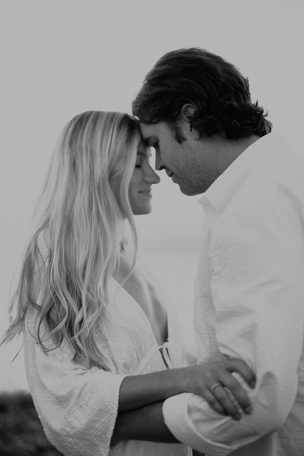 A black and white photo of a couple with foreheads touching and eyes closed, smiling gently, holding each other's hands.