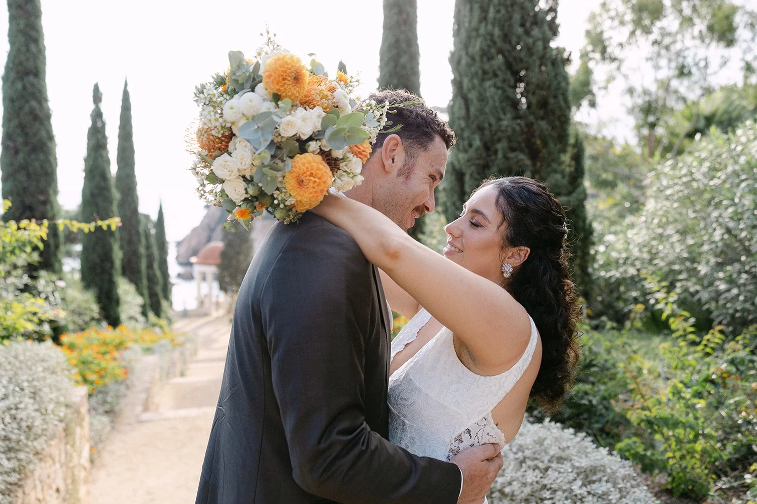 A bride and groom embrace outdoors during their wedding, with the bride holding a large bouquet of yellow and white flowers.