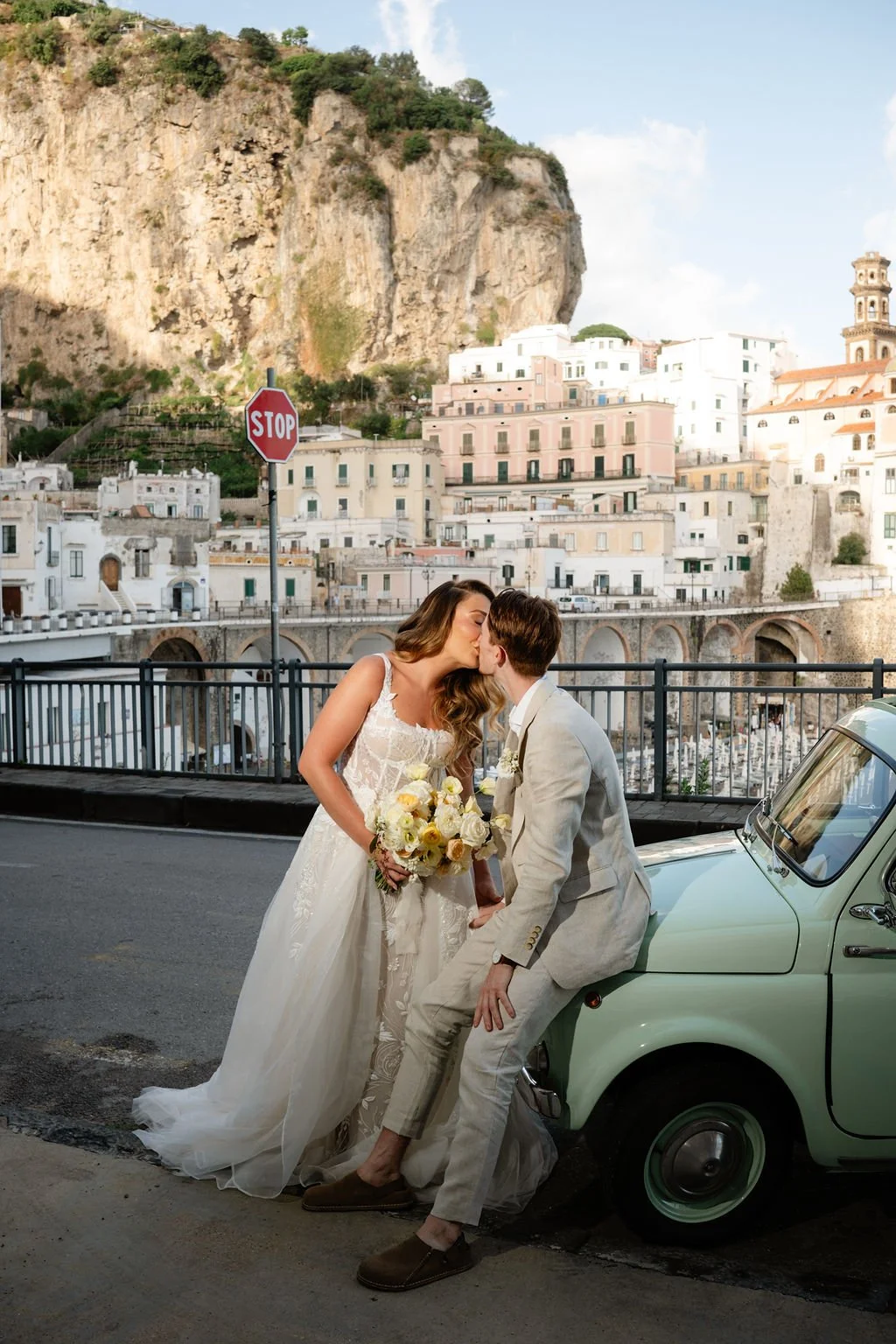 A bride and groom sharing a kiss in front of a vintage car in a scenic European town, with colorful buildings and a rocky hillside in the background.