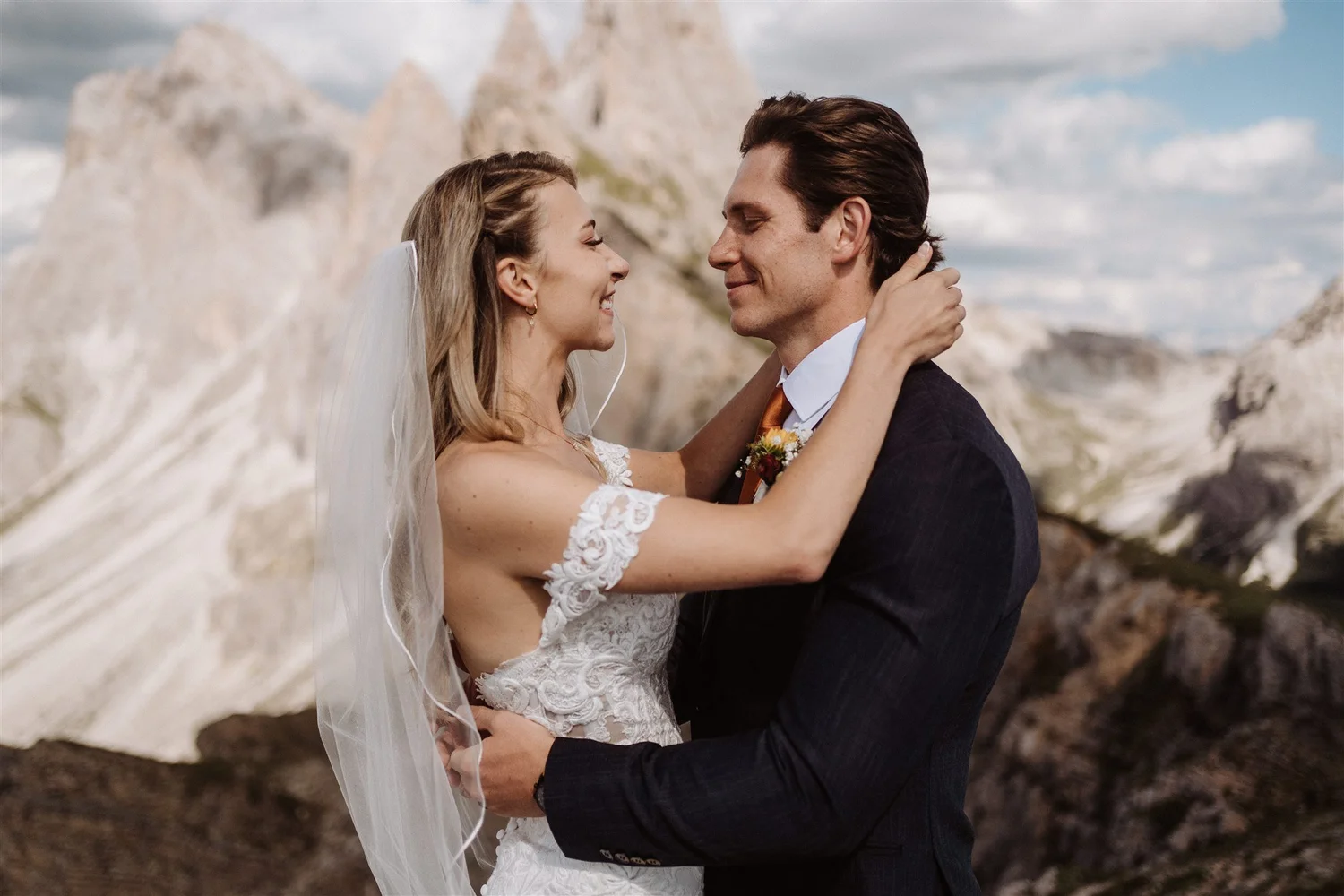 A bride and groom embrace outdoors with mountains in the background.