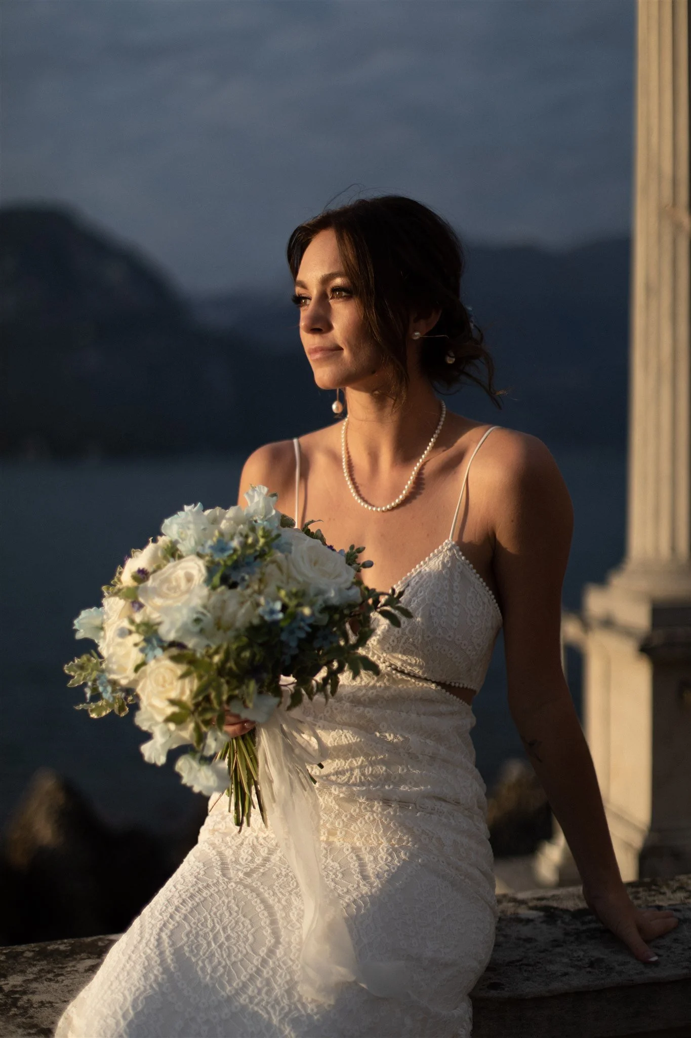 A woman in a white lace dress holding a bouquet of white flowers, sitting outdoors at sunset with mountains and dark clouds in the background.