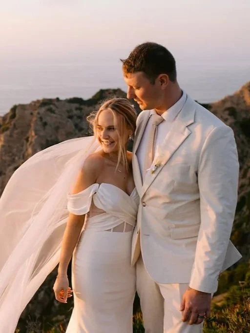 Bride and groom smiling and embracing outdoors during sunset on rocky coastline, dressed in wedding attire.