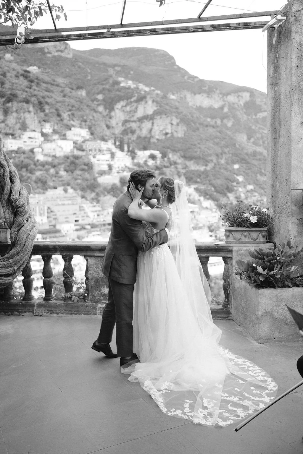 A black and white photo of a bride and groom kissing on a terrace overlooking a mountainous landscape with buildings below. The bride is in a wedding dress with a long lace train, and the groom is in a suit.