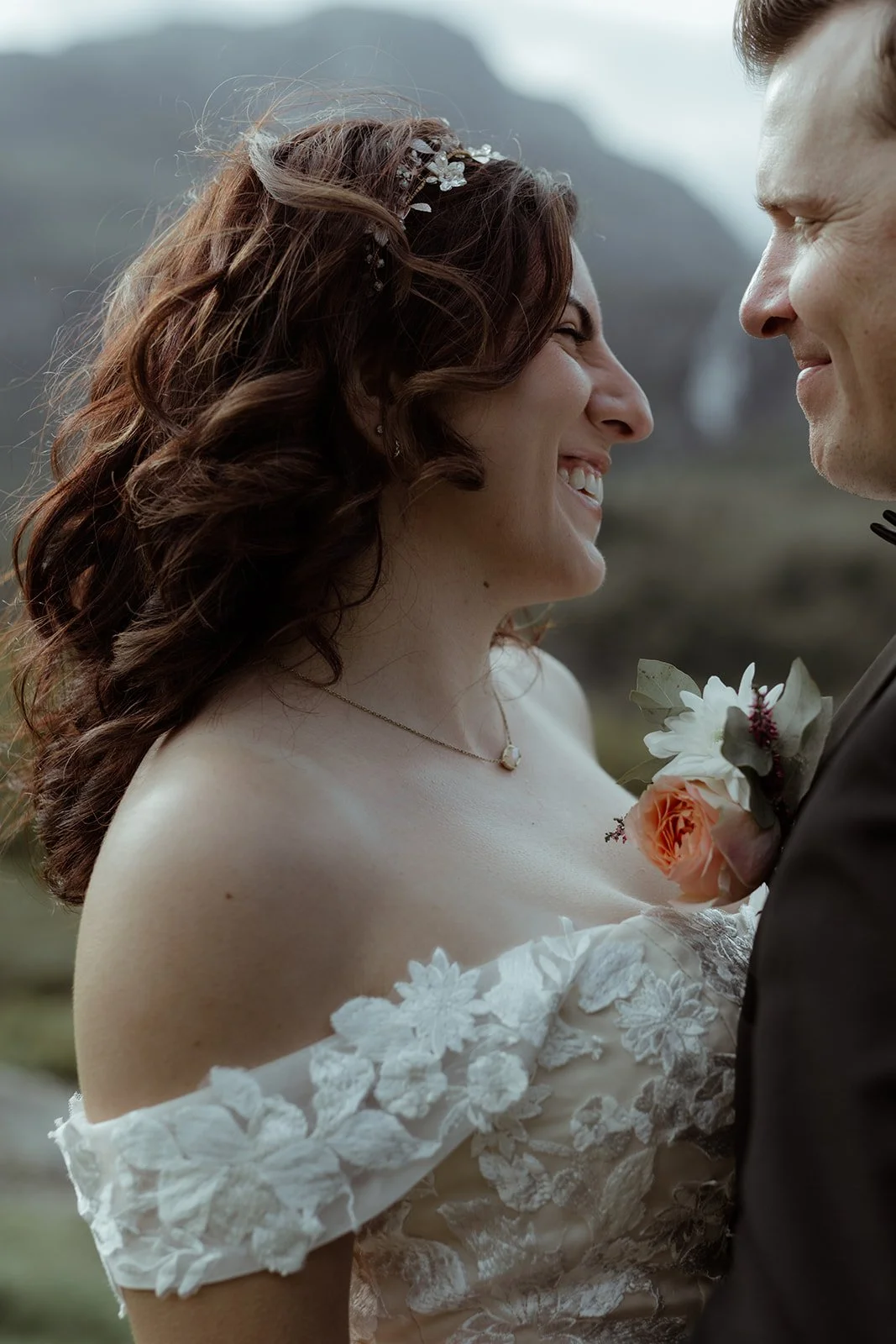 A bride and groom standing close together, smiling and about to kiss outdoors with mountains in the background. The bride has wavy red hair and wears a white lace wedding dress with floral details, a necklace, and a floral hair accessory. The groom i