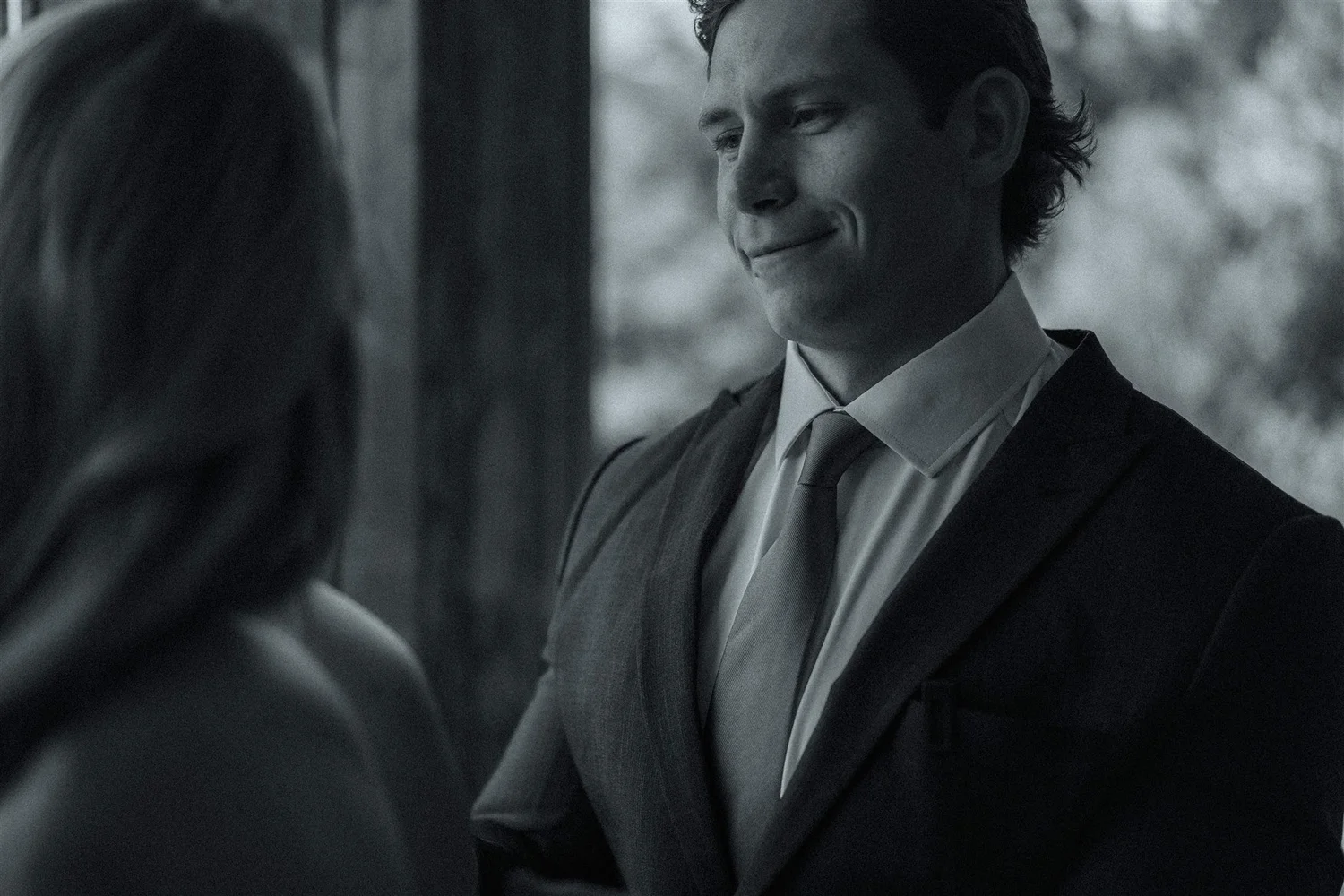 A man in a suit and tie smiling softly at a woman, with trees and outdoor lighting in the background.