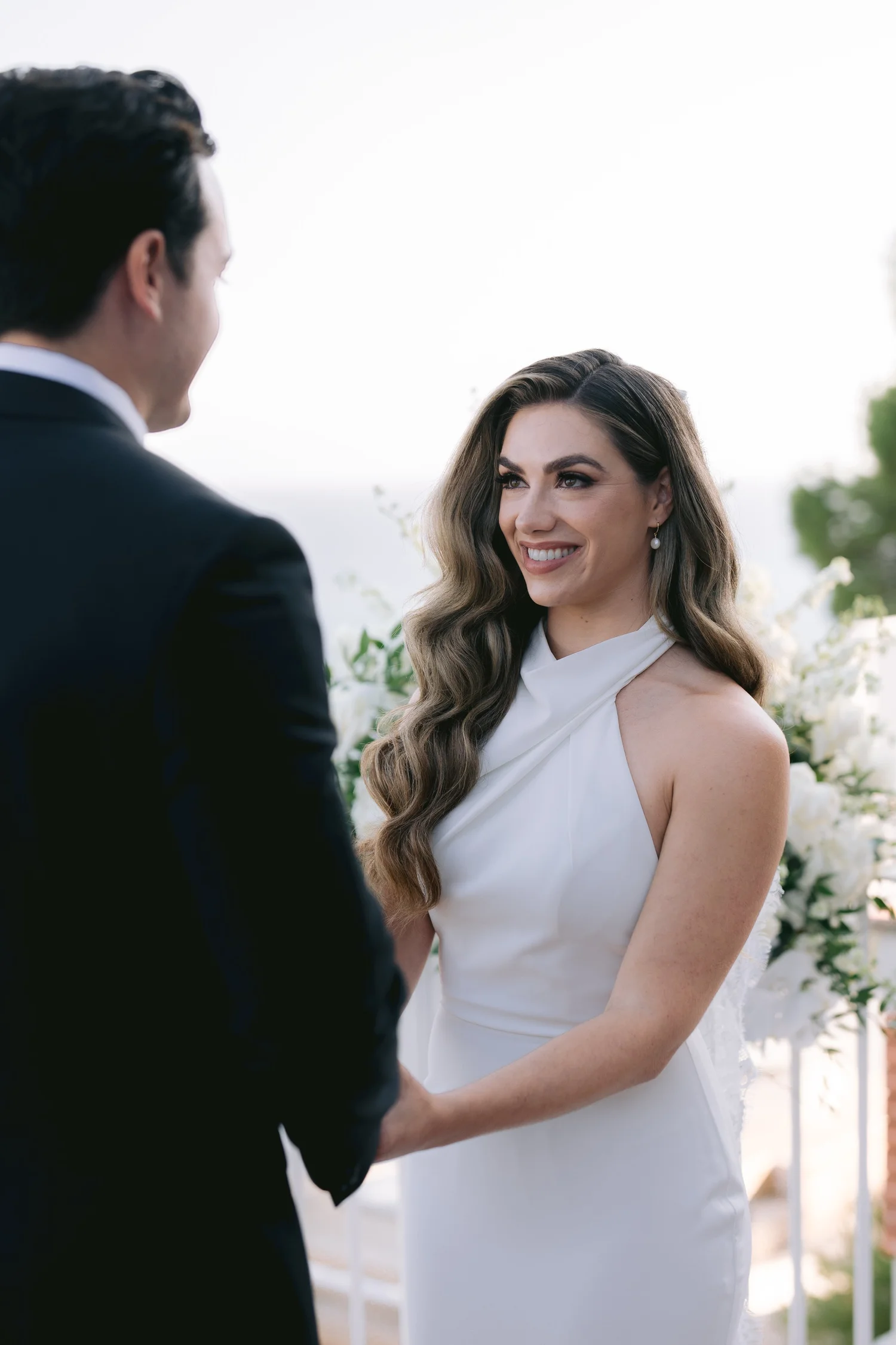 A woman in a white dress happily looks at a man in a black suit during a wedding ceremony outdoors.