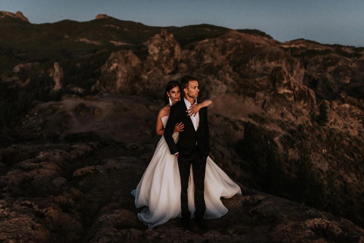 A couple dressed in formal wedding attire standing on rocky terrain with mountains in the background at dusk. The woman is wearing a white wedding gown, and the man is in a black suit and white shirt.