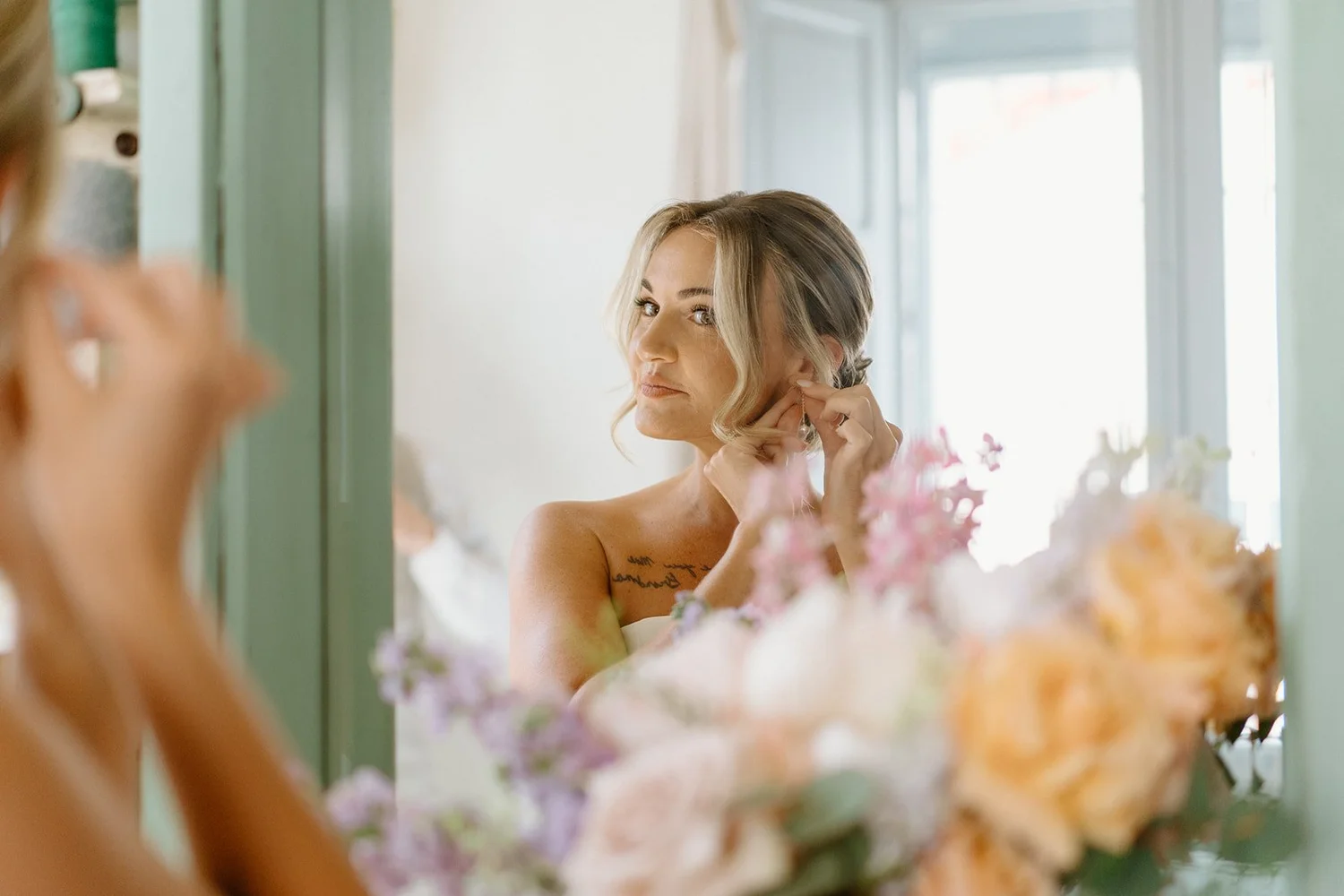 Woman with blonde hair putting on earrings while looking in the mirror, surrounded by soft pastel-colored flowers in a bright room.