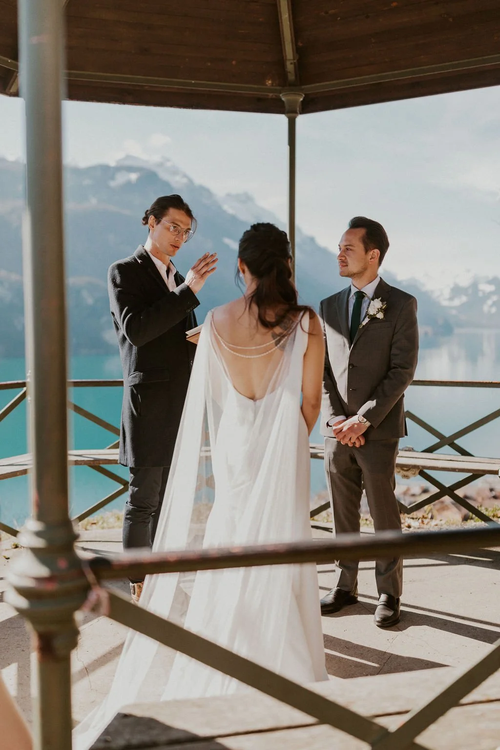 A wedding ceremony taking place outdoors with mountains and a lake in the background, featuring a bride and groom facing an officiant under a wooden gazebo.