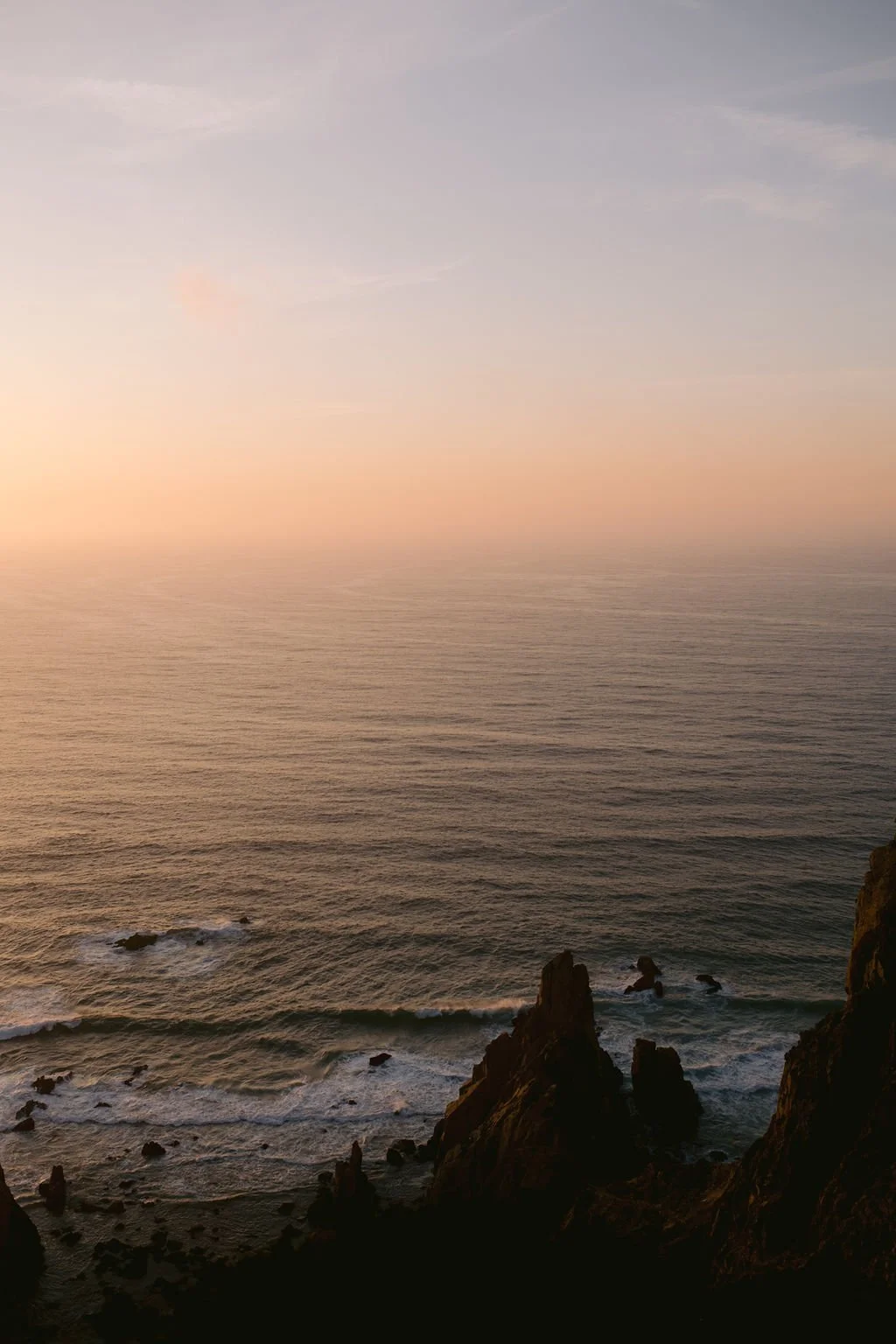 View of the ocean at sunset with rocky cliffs in the foreground and a pastel-colored sky.