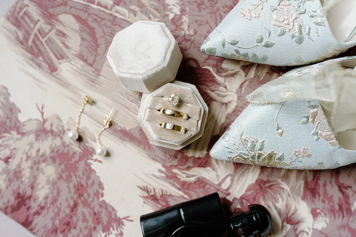 Open velvet jewelry box with a diamond engagement ring and a wedding band, surrounded by pearl earrings, floral fabric, and lipstick on a pink patterned surface.