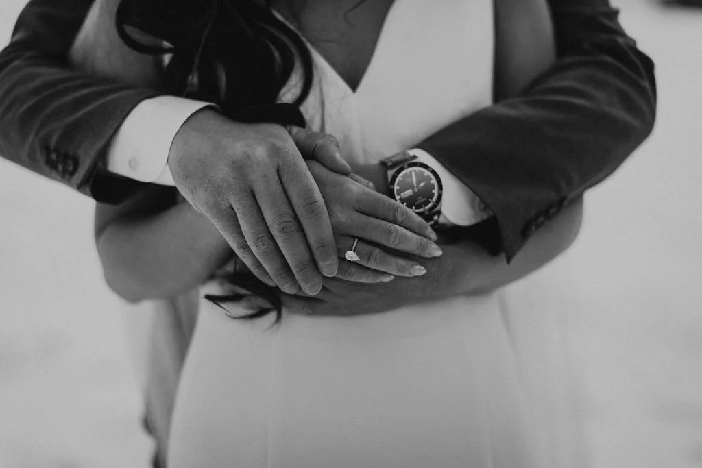 A couple's hands showing an engagement ring, with one person wearing a watch and the other a suit jacket, in a close-up black and white photo.
