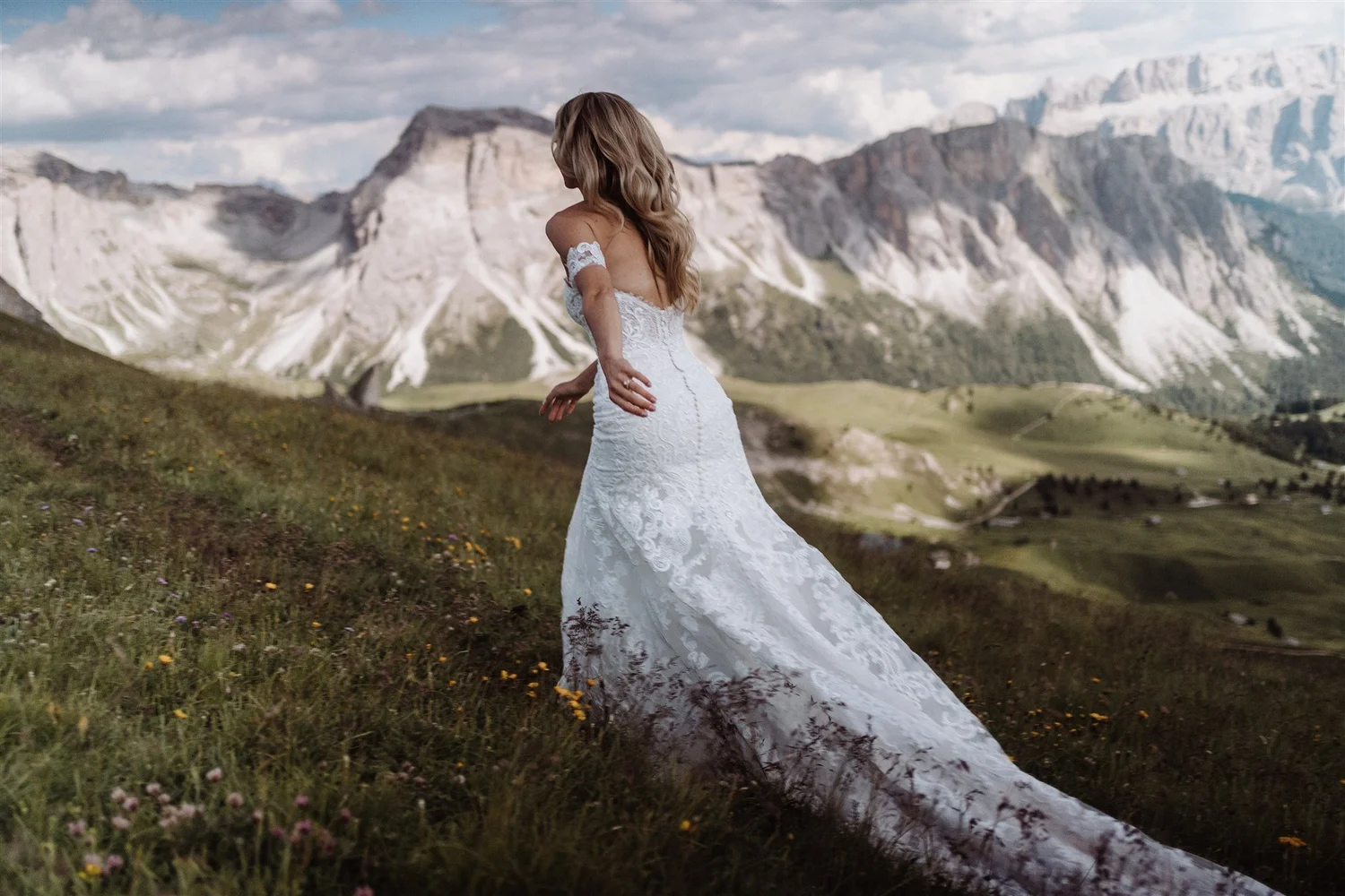 A woman in a white wedding dress standing on a grassy hillside with snow-capped mountains and a cloudy sky in the background.