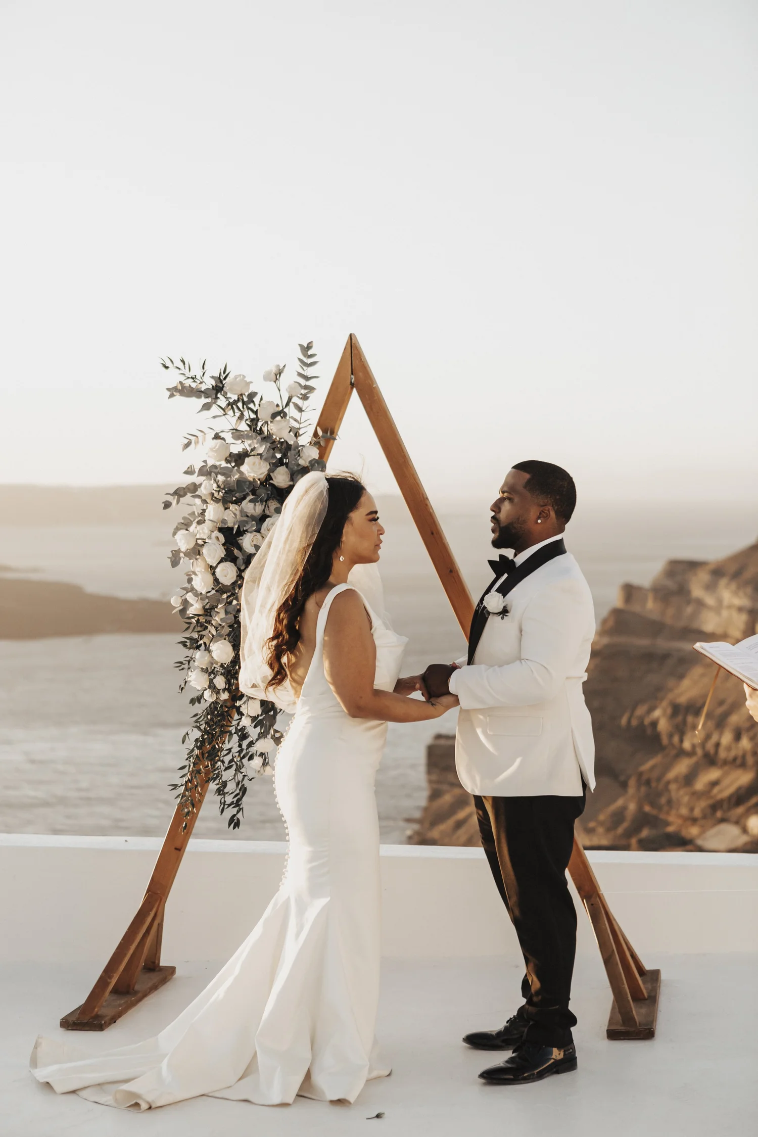 A bride in a white wedding dress and veil holding hands and looking at a groom in a white tuxedo jacket with black bow tie and black pants, standing on a sandy beach at sunset with rocky cliffs in the background. They are under a wooden wedding arch
