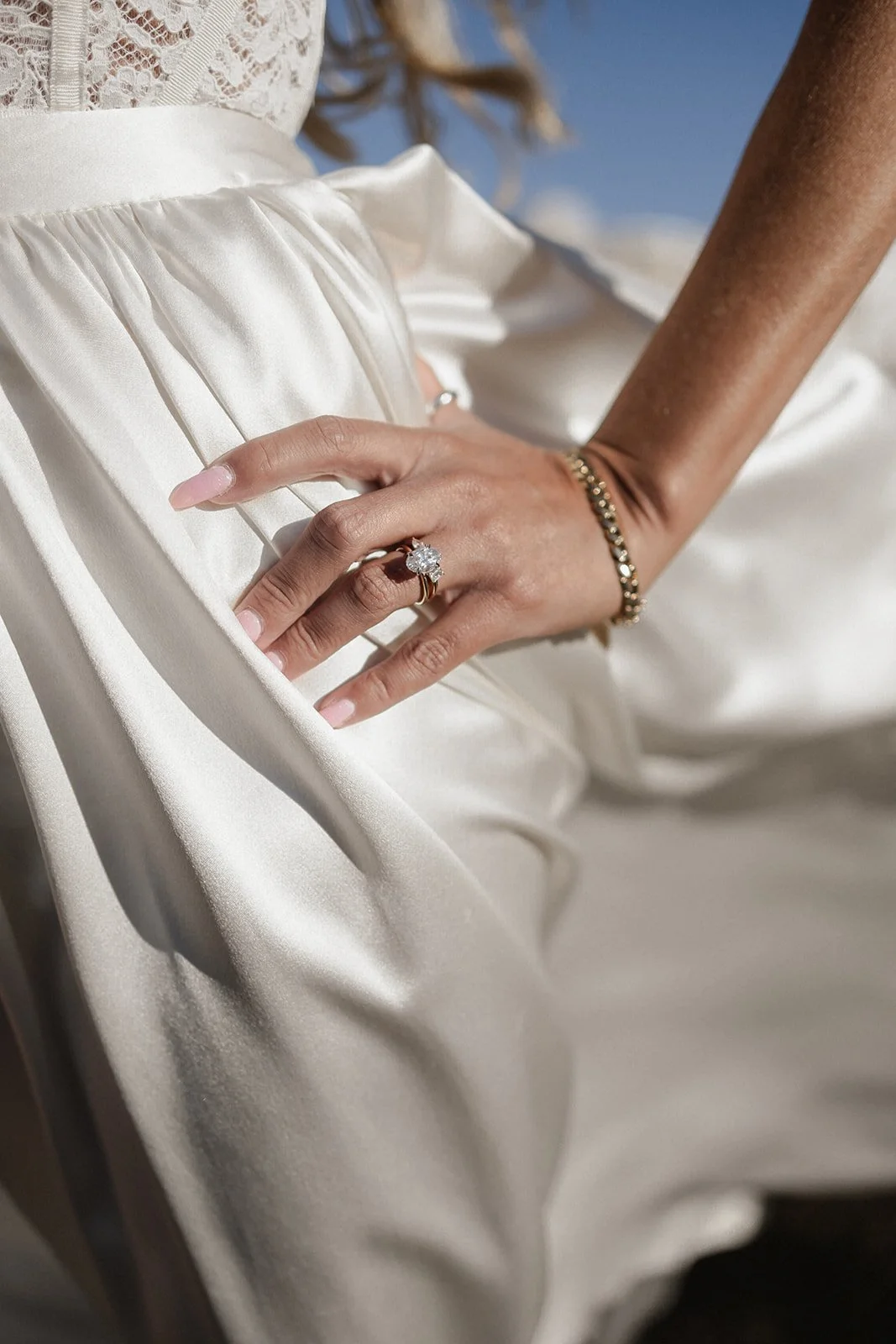 Close-up of a woman's hand with a large diamond engagement ring and a simple gold band, resting on her waist, with a lace dress and satin fabric in the background.