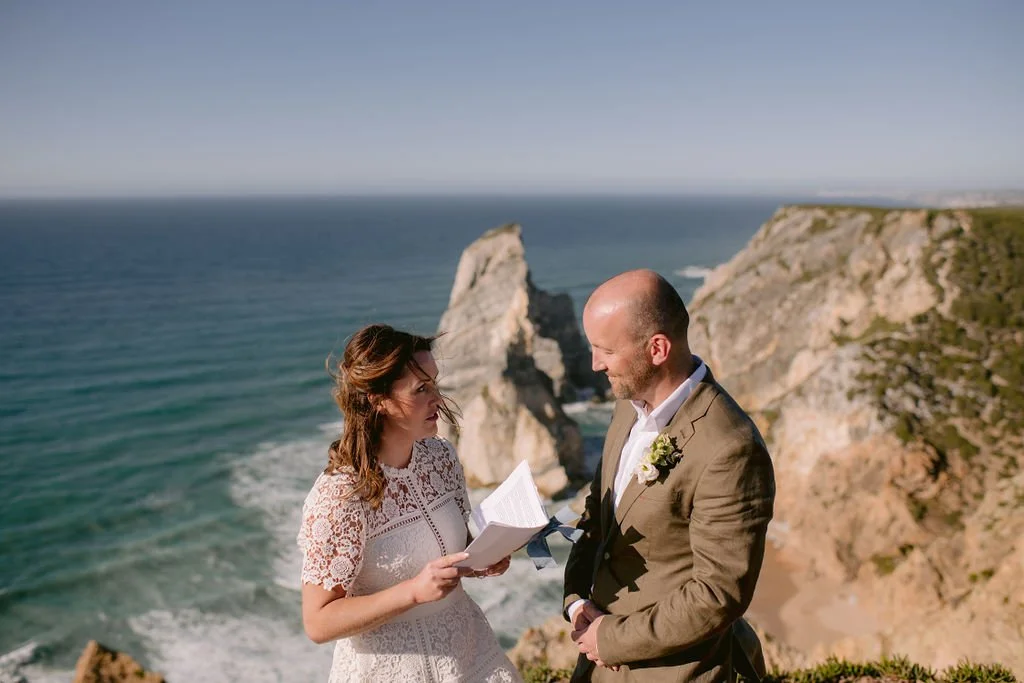 A couple getting married on a cliff overlooking the ocean, with the bride holding a book and the groom looking at her, both dressed in wedding attire.
