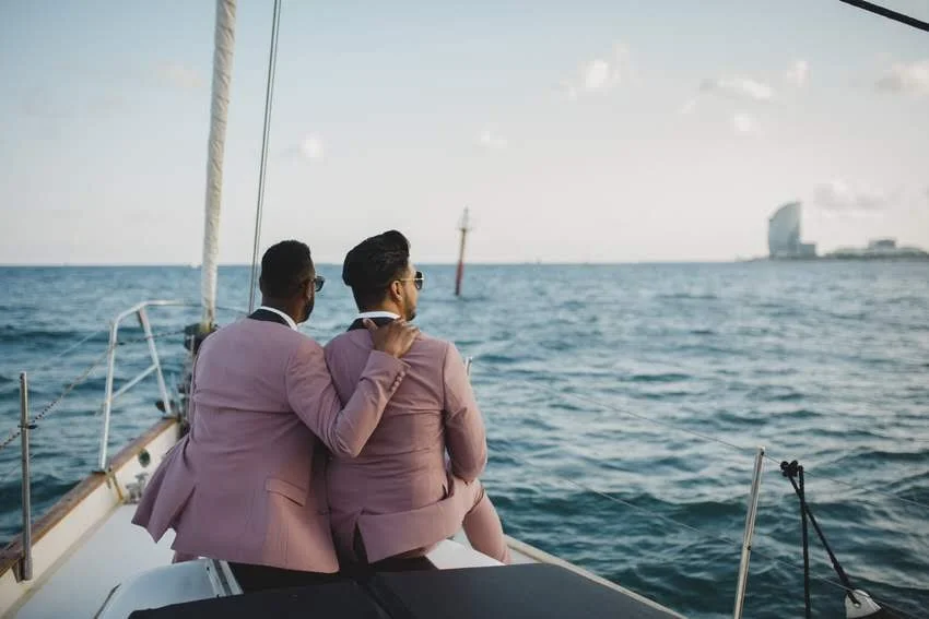 Two men in pink suits sitting on a yacht, looking out at the ocean with a city skyline in the distance.