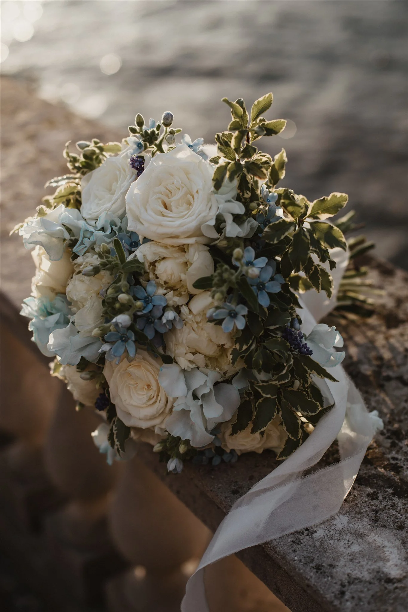 A bouquet of white roses, blue delphiniums, and greenery wrapped in white ribbon, resting on a stone surface near water with sunlight reflecting off the water.