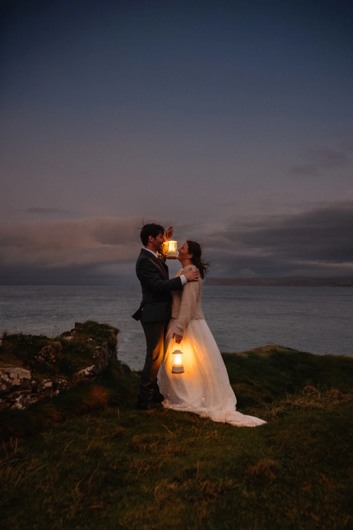 A couple standing outdoors at night, holding lanterns, with the woman wearing a wedding dress and the man in a suit, by the water, with a dark sky and clouds in the background.