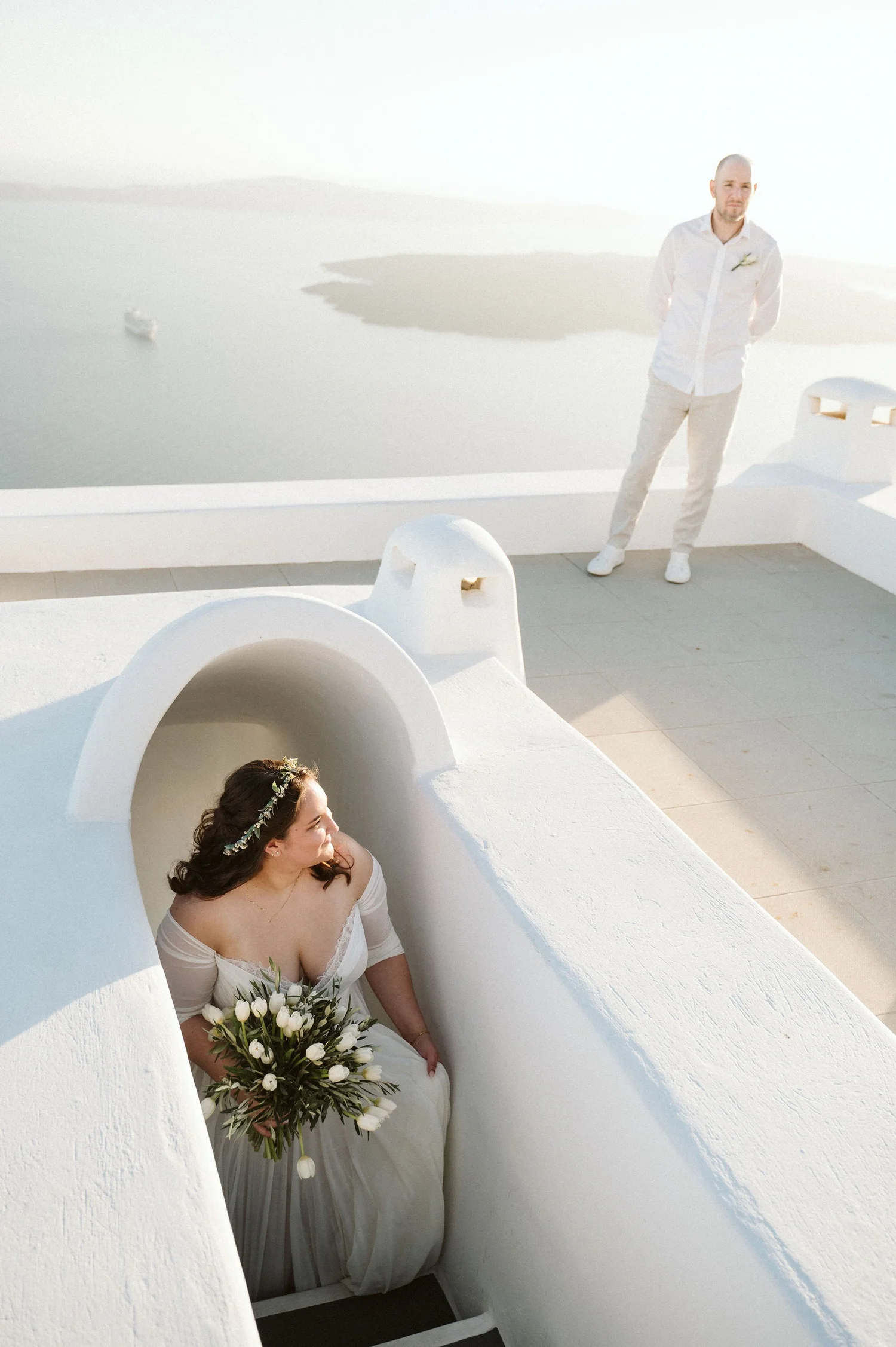 A bride sitting in a white, curved architectural niche holding a bouquet of white flowers with greenery, in a white dress with off-the-shoulder sleeves, looking to her left, with a man standing outside on a white smooth platform, dressed in beige pan