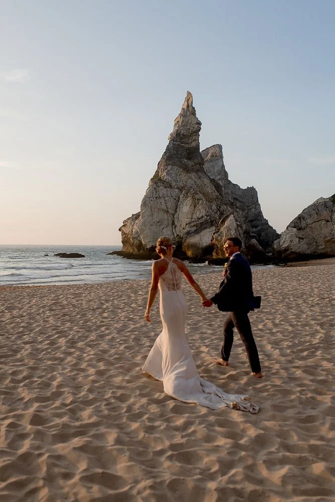 A bride and groom holding hands walk on a sandy beach with a large rocky formation in the background during sunset.