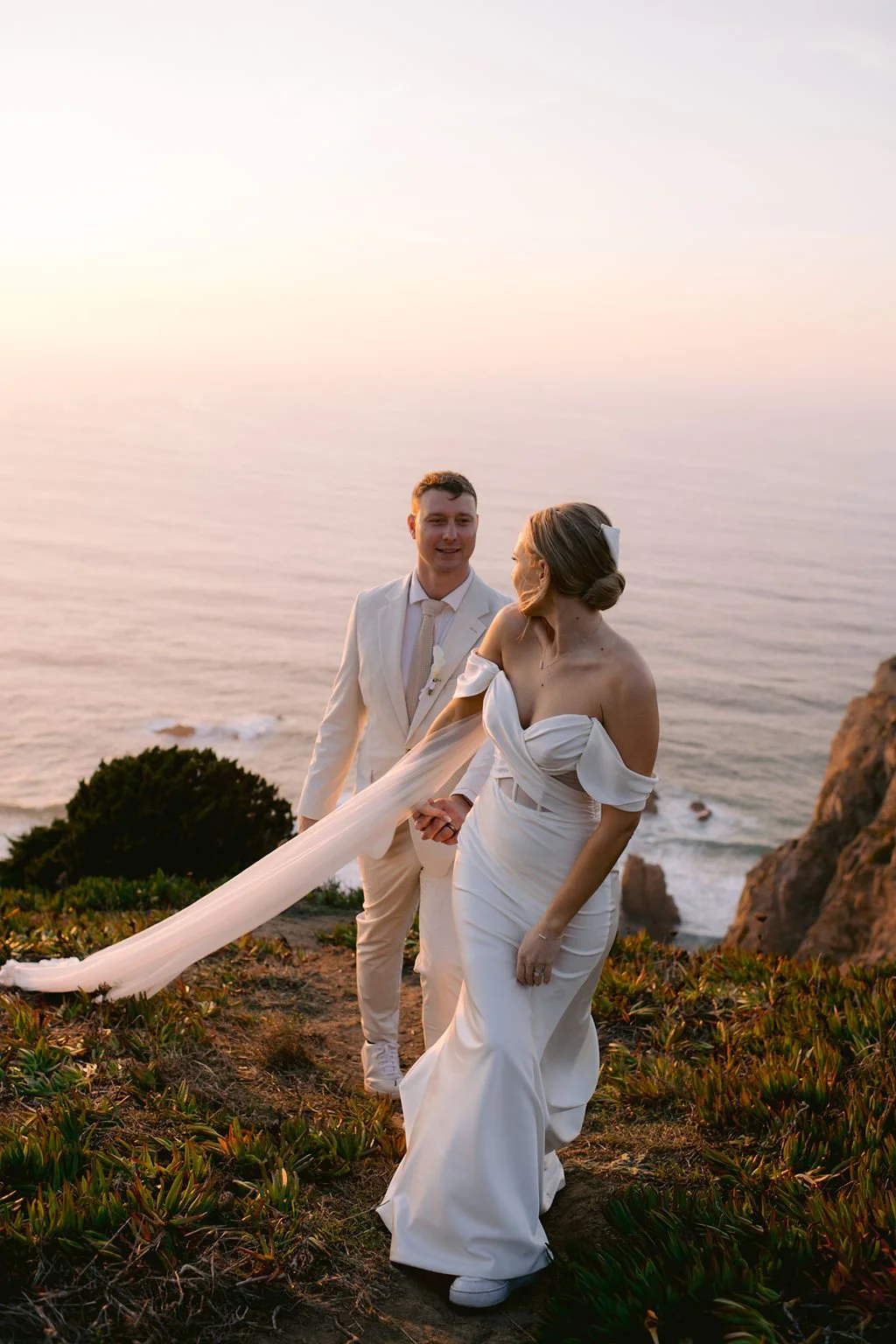 A bride and groom hold hands on a cliffside overlooking the ocean during sunset.