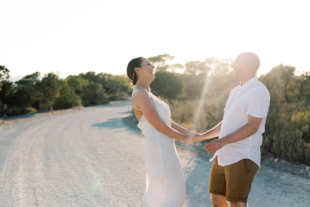 A couple holding hands and smiling at each other on a dirt road with trees in the background during sunset.