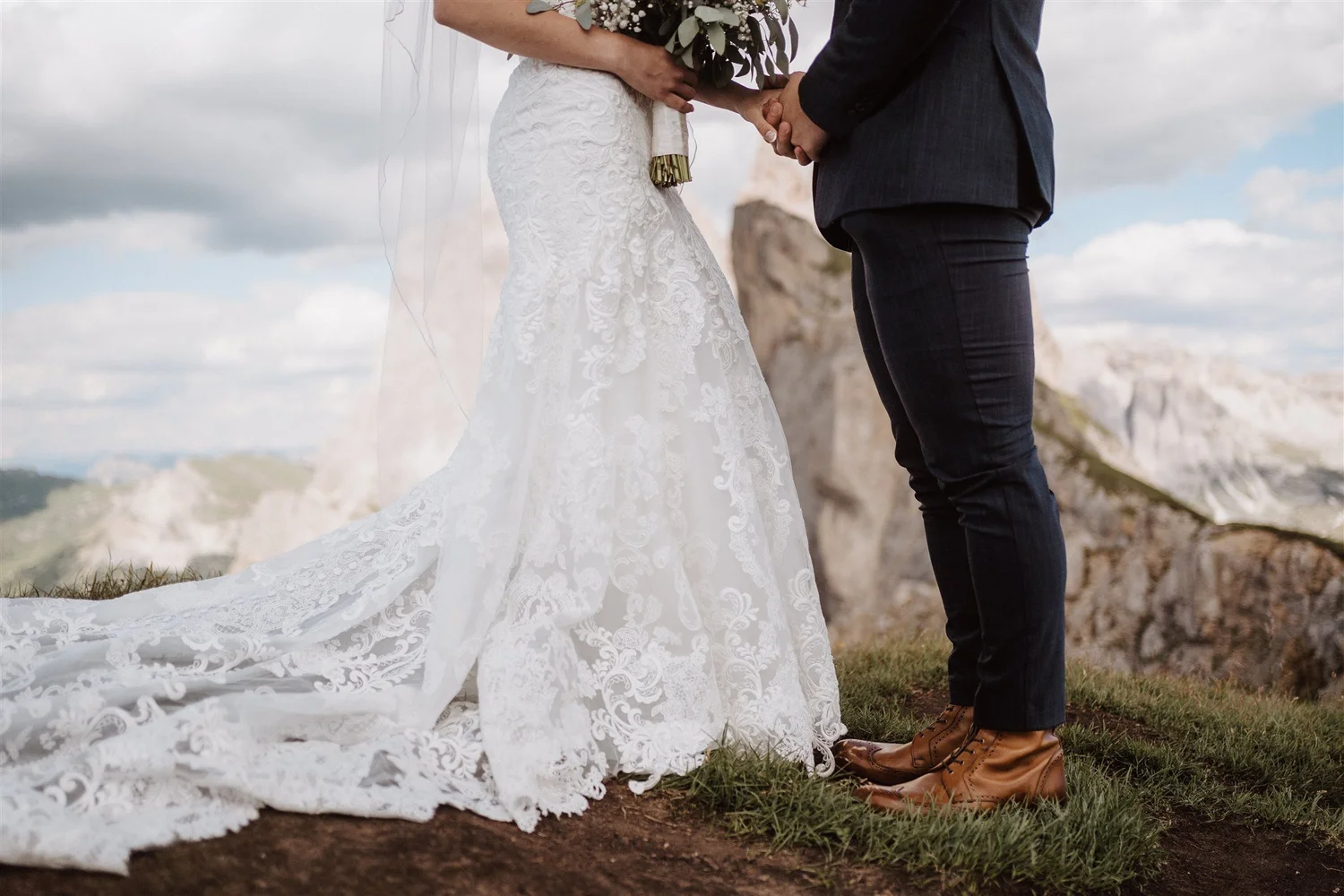 A couple holding hands during their outdoor wedding ceremony, with the bride in a white lace gown and the groom in a dark suit with brown shoes, standing on grass with mountains and cloudy sky in the background.