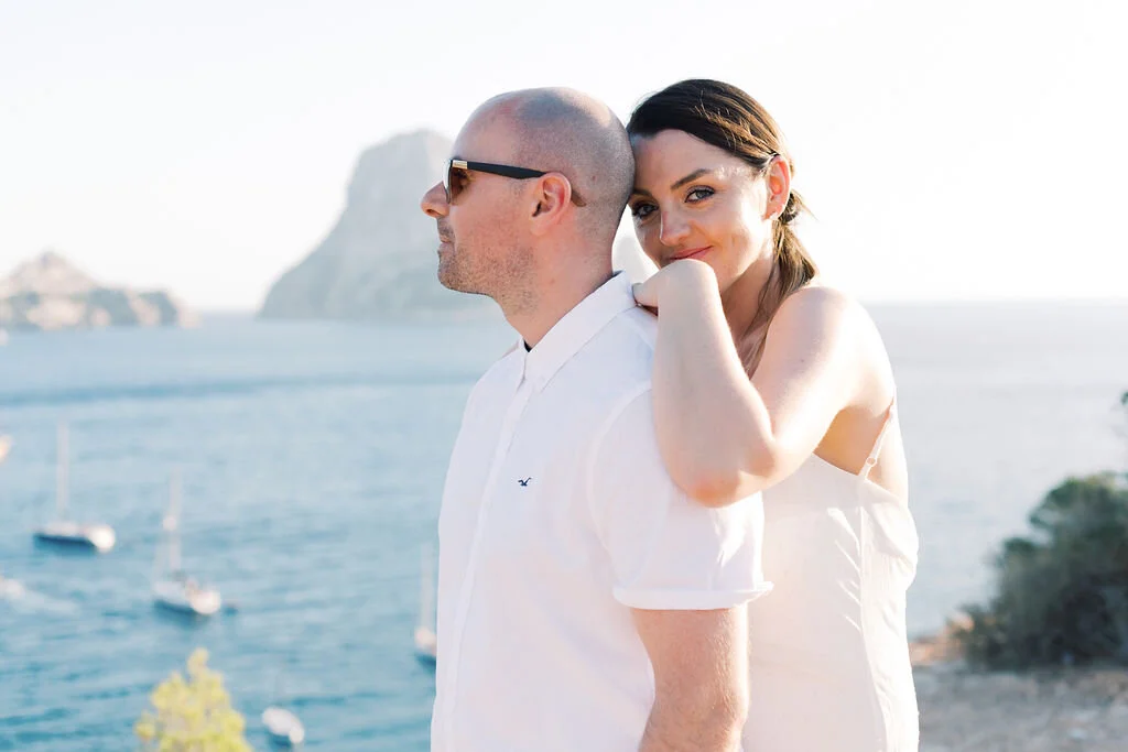A woman with dark hair hugging a bald man with sunglasses near the ocean with boats and cliffs in the background.