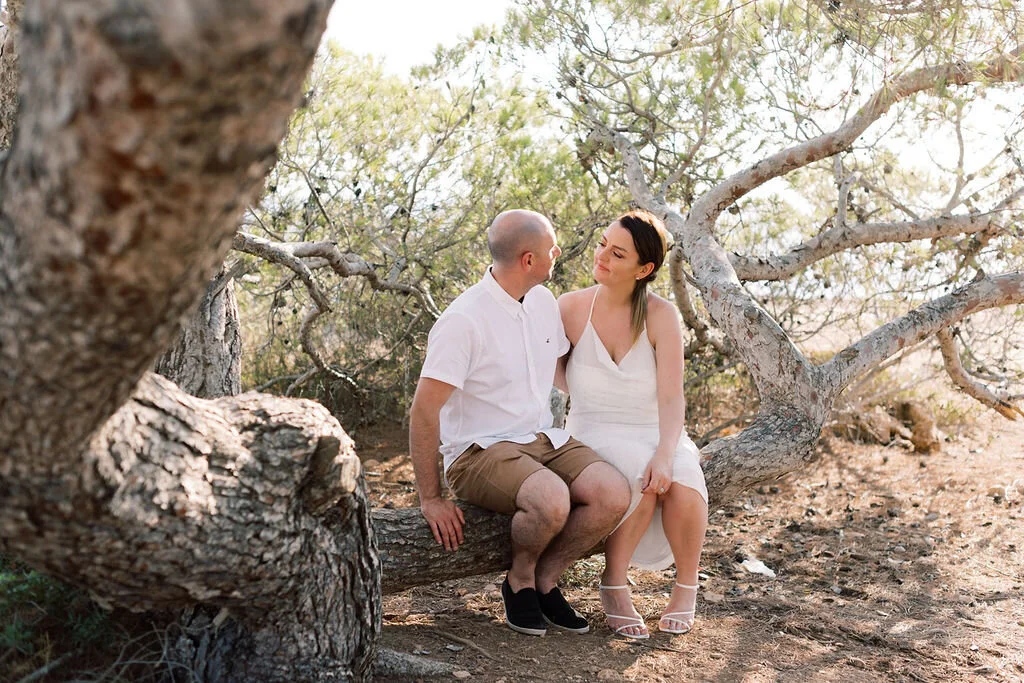 A couple sitting on a tree branch in a wooded area, looking at each other. The man is bald, wearing a white shirt and shorts. The woman has dark hair, wearing a white dress and heels.