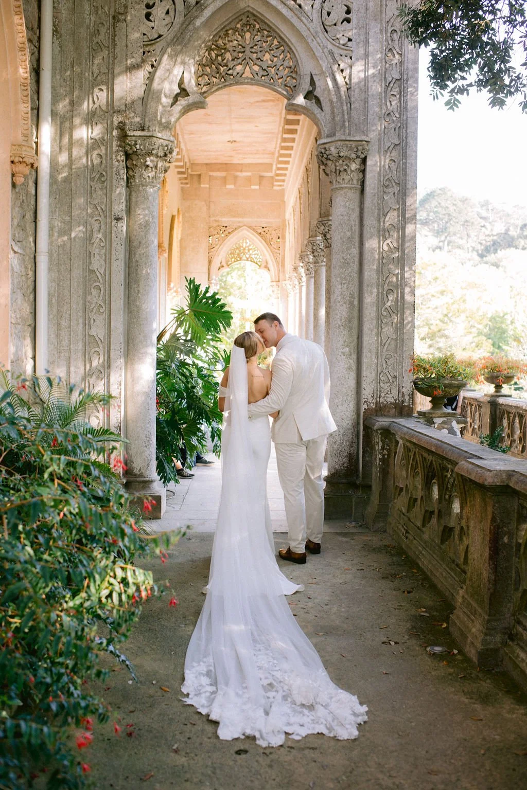 A bride and groom share a kiss under an ornate stone archway during their wedding, surrounded by lush greenery and historic architecture.