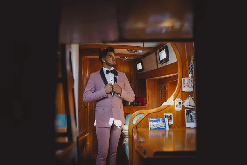 A man in a pink suit and black bow tie adjusting his jacket inside a wooden-paneled room, possibly on a boat or yacht, with photos and small screens on the wall.