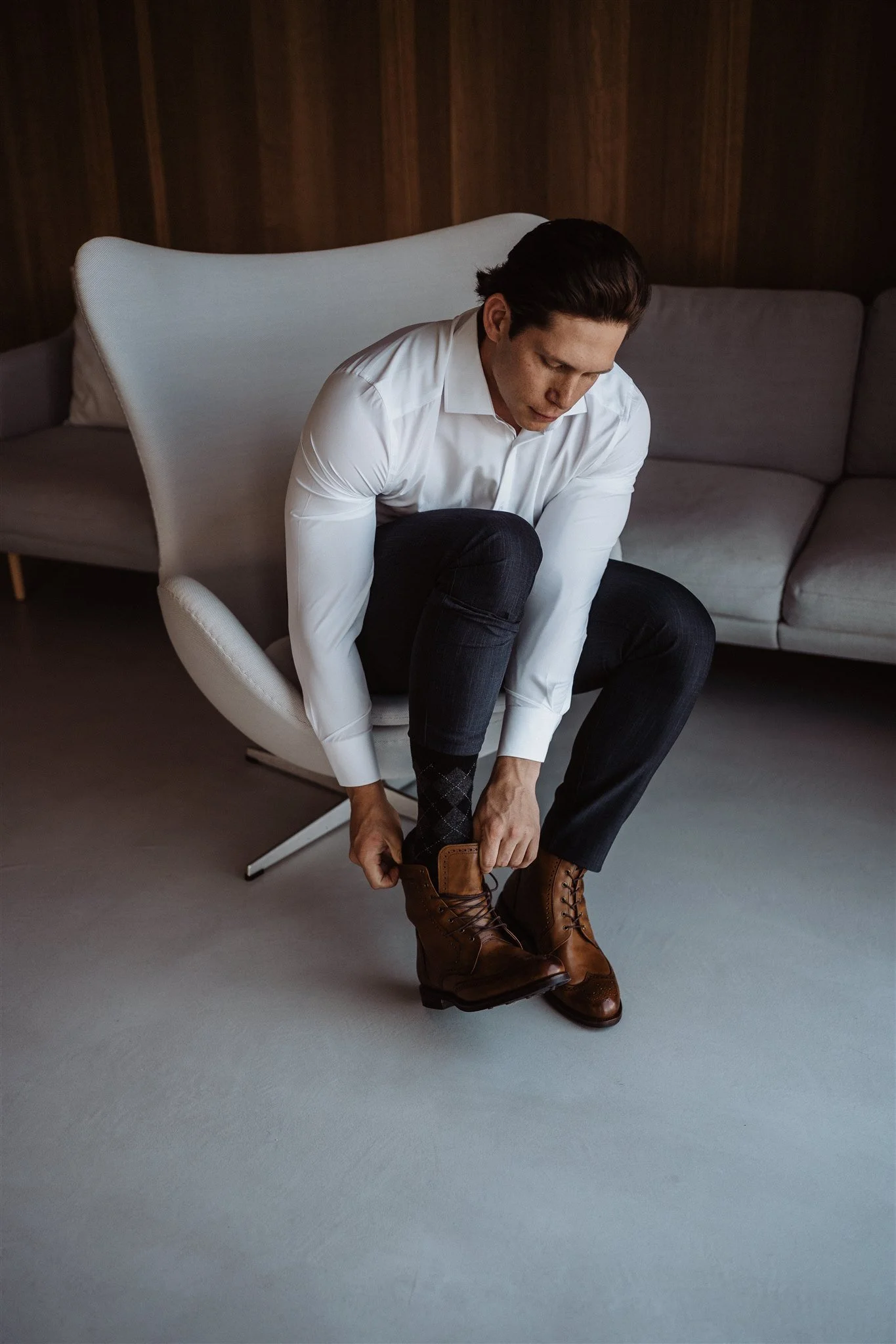 A man in a white dress shirt and black pants is sitting in a modern white chair, tying the laces on his brown leather boots.