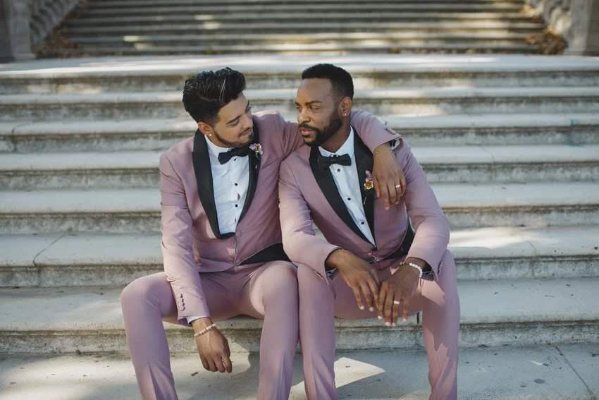 Two men wearing pink tuxedos with black lapels, sitting on outdoor steps. One has an arm around the other's shoulder, and they are looking at each other affectionately.