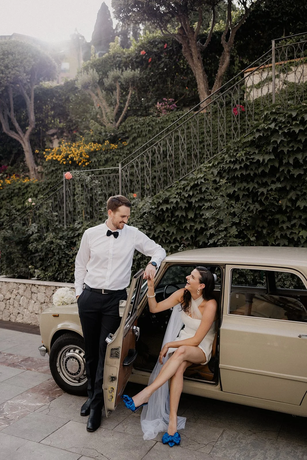 A man and a woman dressed in formal attire, smiling and holding hands near a vintage beige car with an outdoor, lush garden background.