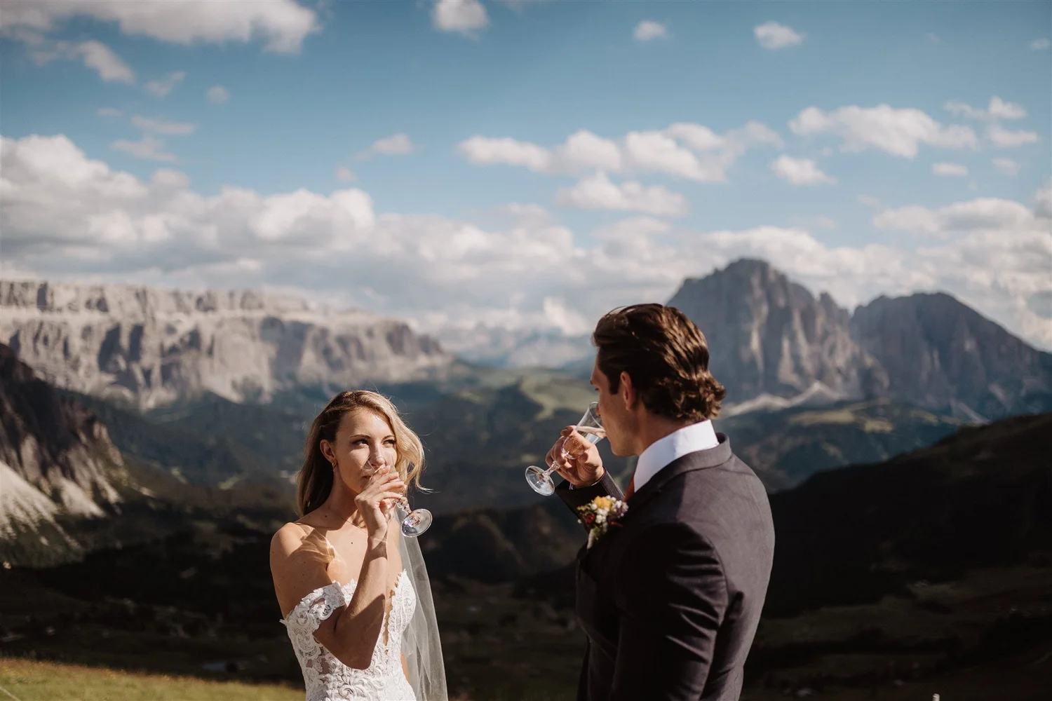 A bride and groom are outdoors in a mountainous landscape, toasting with glasses of champagne.