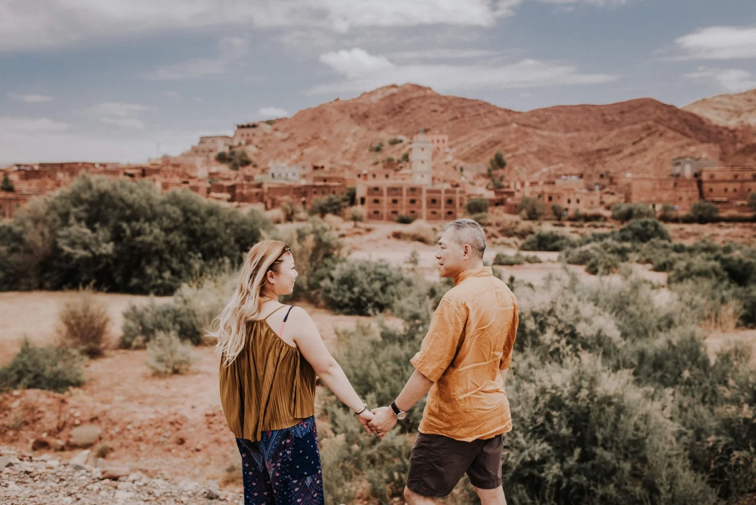 A couple holding hands and looking at each other in a desert landscape with red rock formations and sparse vegetation.