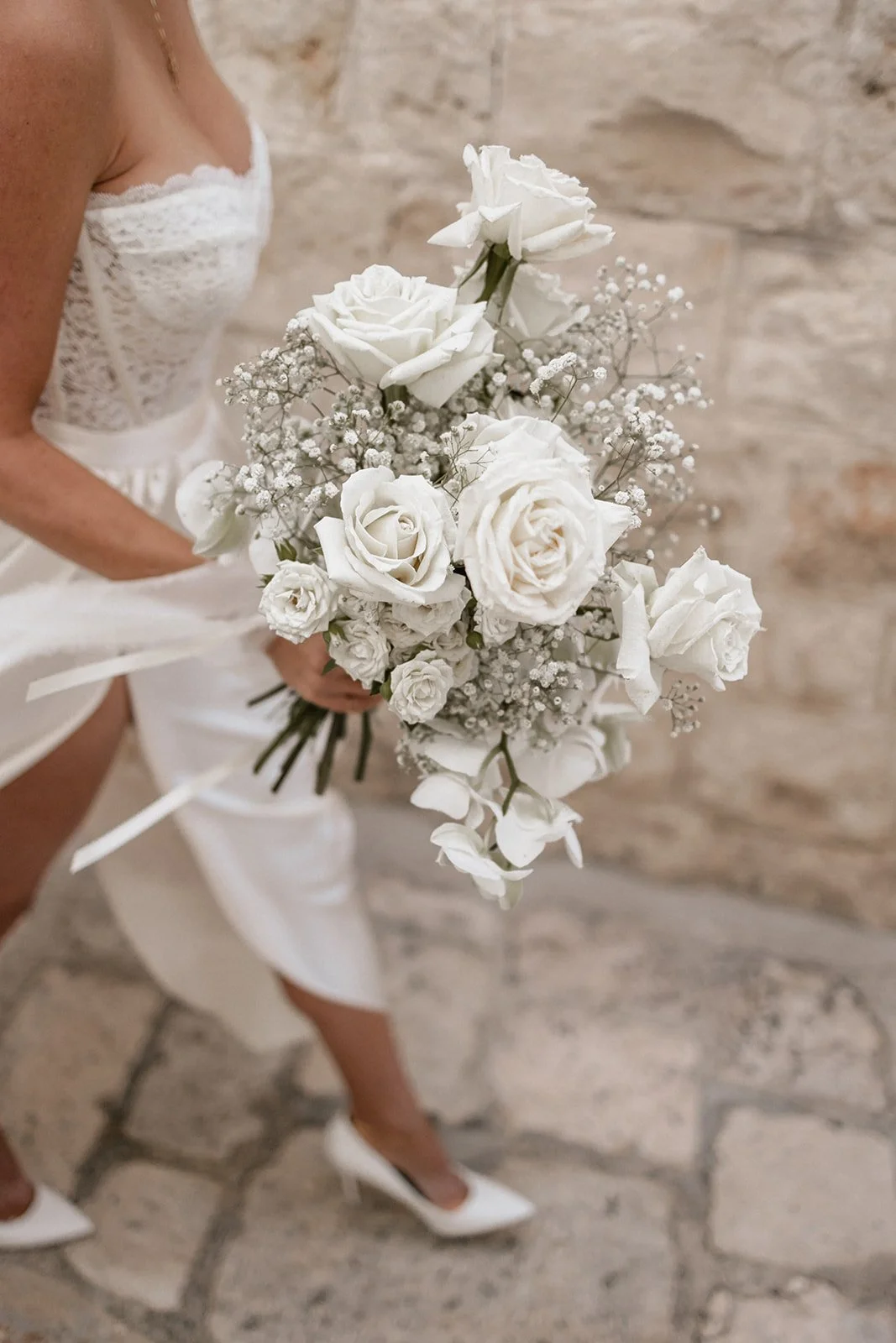 A woman in a white lace dress holding a bouquet of white roses and baby's breath, standing on a stone floor against a stone wall background.