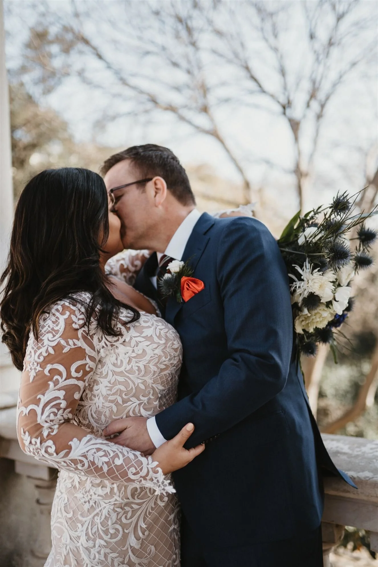 A couple sharing a kiss at their wedding, with trees in the background and the bride holding a bouquet of flowers.
