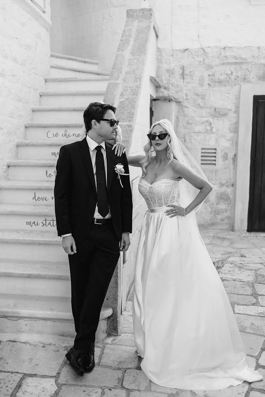 Black and white photo of a man and woman in wedding attire standing against a staircase. The man is wearing a black suit, white shirt, black tie, and dark sunglasses. The woman is wearing a strapless wedding gown, sunglasses, a veil, and has long blo